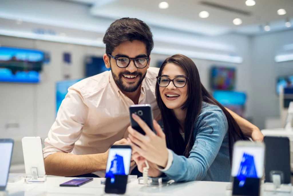 Happy couple wearing glasses smiling while looking at cell phone with in store promotions display phones in store